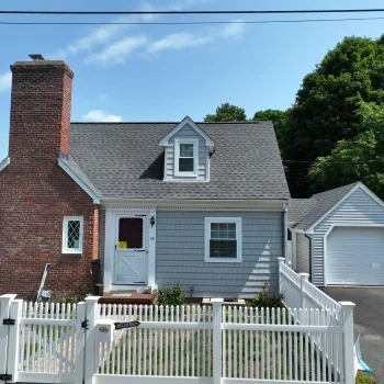 Front exterior view of a small residential home with a brick chimney, vinyl siding and a shingled slanted roof with a gated front yard and driveway